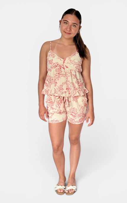 A woman modeling a two-piece matching set featuring a ruffled camisole top and shorts in a pink toile de Jouy pattern on a cream background, set against a plain light gray studio background.