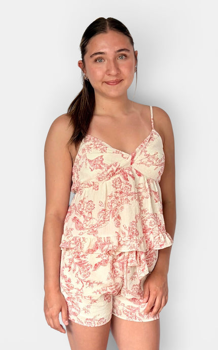 A young woman modeling a two-piece camisole and shorts set with a pink toile de Jouy pattern on a cream background, set against a plain light gray studio background.