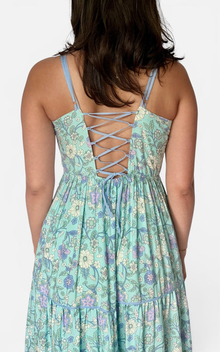 A woman shown from the back wearing a sleeveless tiered dress with thin shoulder straps, a floral print in mint green, purple, and cream, and a lace-up open back.