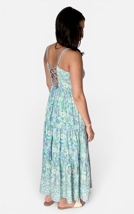 A woman shown from the back wearing a light blue maxi dress with a white and green floral pattern and lace-up back straps, standing against a plain light gray background.