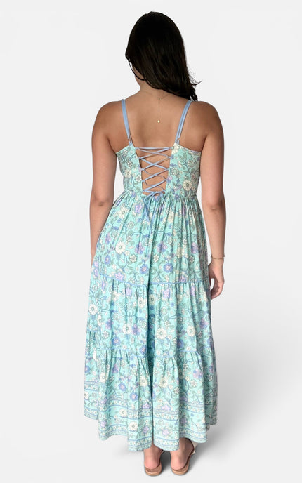 A woman viewed from the back wearing a light aqua maxi dress with a floral print and lace-up back detail, set against a plain light gray studio background.