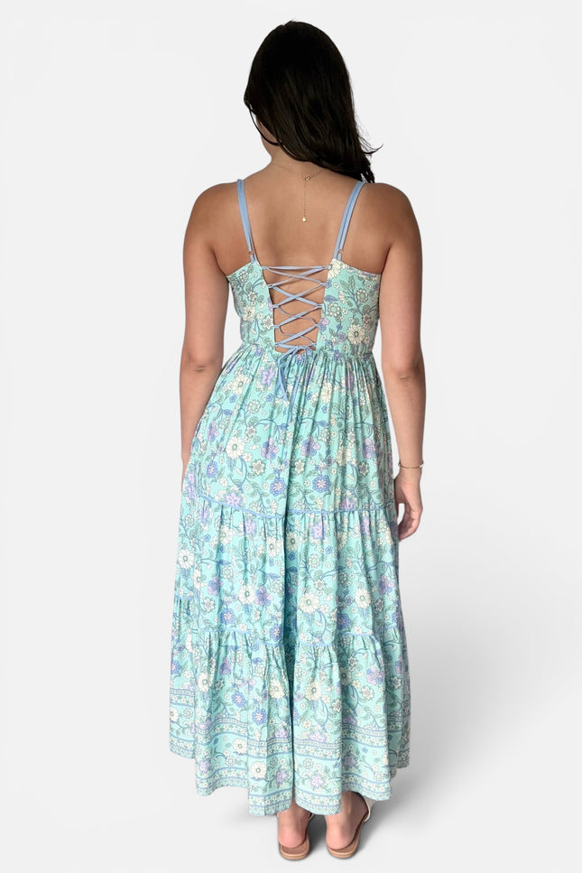 A woman viewed from the back wearing a light aqua maxi dress with a floral print and lace-up back detail, set against a plain light gray studio background.