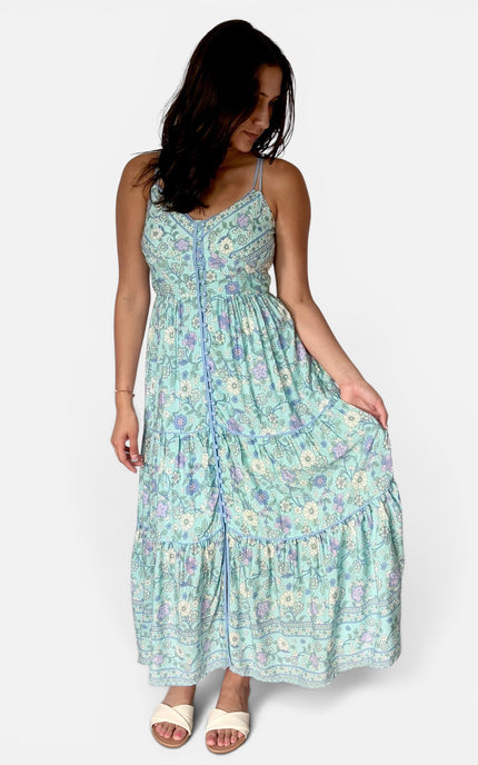A woman modeling a light blue and purple floral print maxi dress with spaghetti straps and a tiered skirt, paired with white slide sandals, against a plain light gray background.