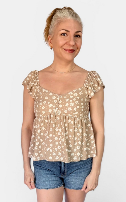 A woman modeling a beige short-sleeve peplum top with a white floral pattern, paired with denim shorts, against a plain light gray background.