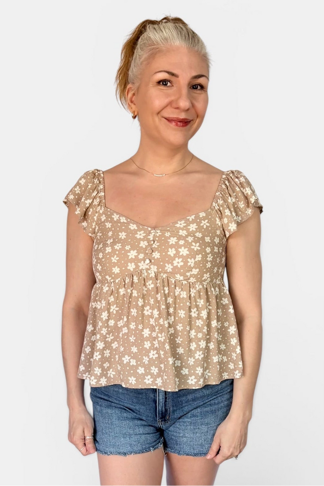 A woman modeling a beige short-sleeve peplum top with a white floral pattern, paired with denim shorts, against a plain light gray background.