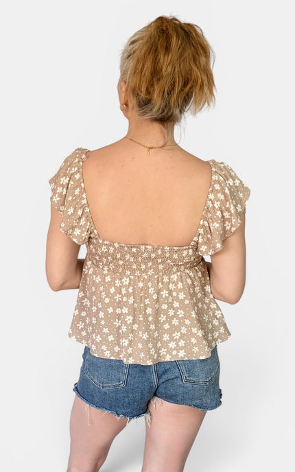 A woman shown from the back wearing a beige floral ruffled top and denim shorts against a light gray studio background.