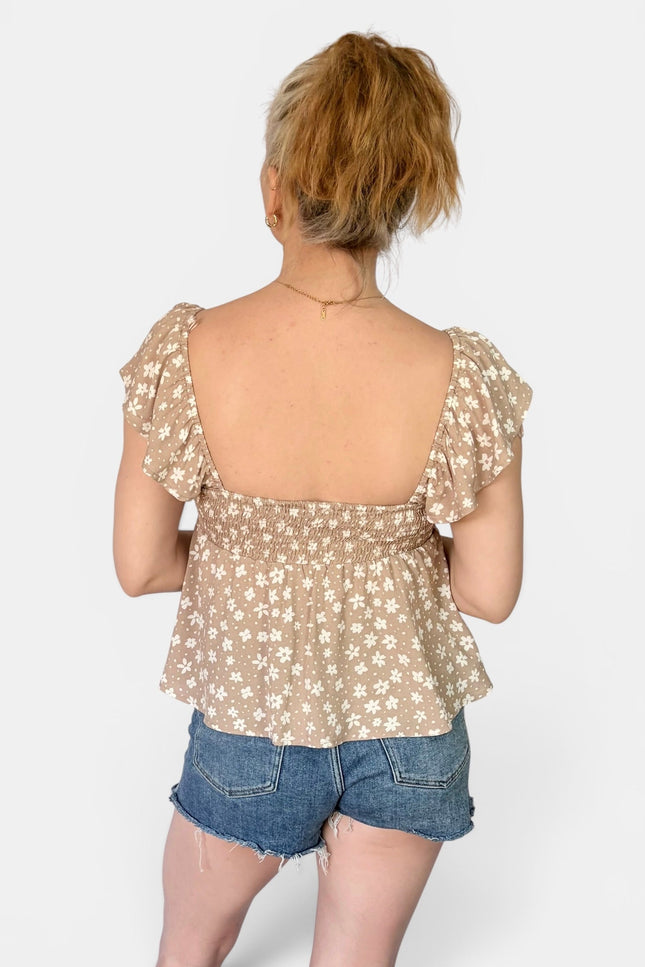 A woman shown from the back wearing a beige floral ruffled top and denim shorts against a light gray studio background.
