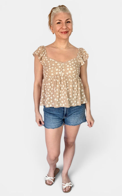 A woman modeling a beige short-sleeve top with a white floral pattern, paired with denim shorts and white sandals, against a light gray background.