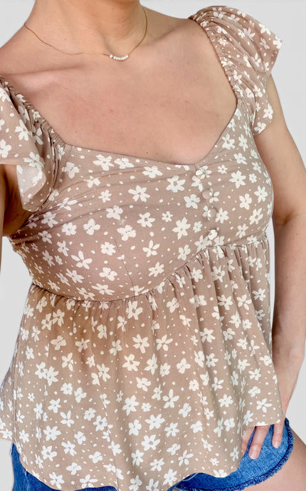 A close-up of a woman wearing a taupe-brown off-the-shoulder top with a white floral print and ruffled sleeves, paired with blue denim shorts.