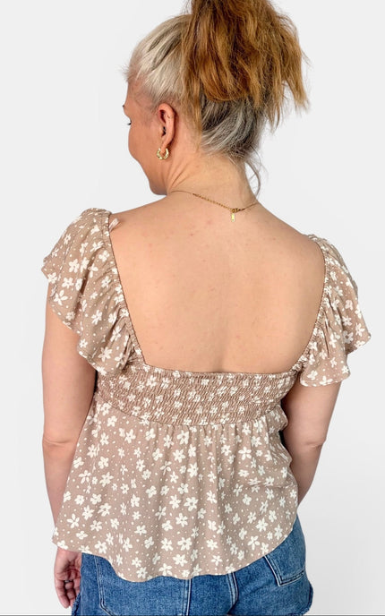 A woman viewed from the back wearing a beige puff-sleeve top with a white floral pattern and a smocked waist, paired with blue jeans against a light gray background.