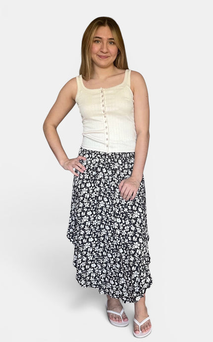 A woman modeling a sleeveless cream top and a black midi skirt with a white floral pattern, paired with white sandals, against a light gray studio background.