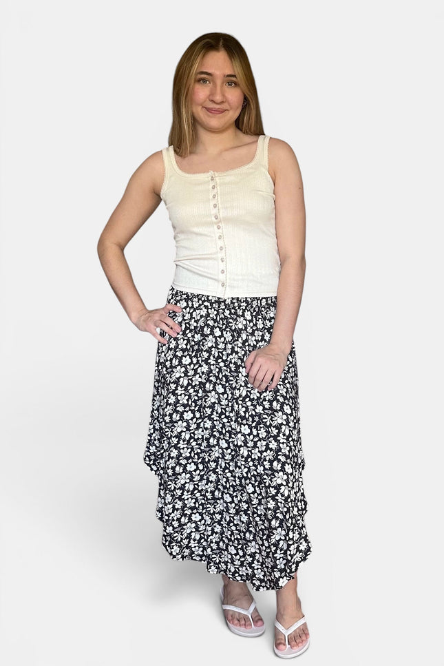 A woman modeling a sleeveless cream top and a black midi skirt with a white floral pattern, paired with white sandals, against a light gray studio background.