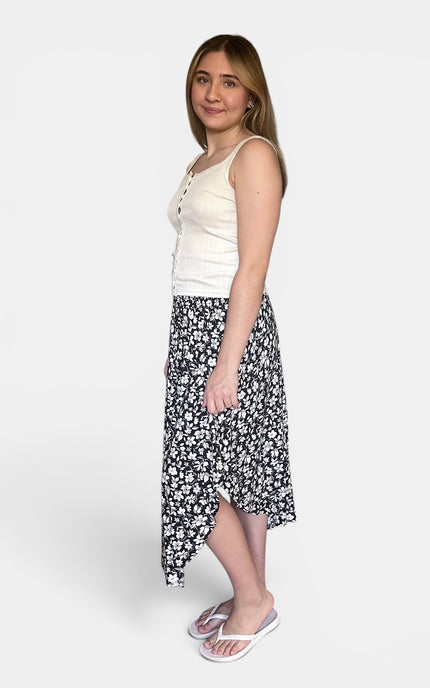 A woman modeling a black and white floral midi skirt paired with a cream tank top and white flip-flops, standing against a light gray studio background.