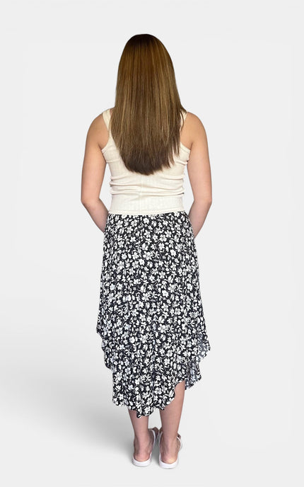 A woman viewed from behind wearing a sleeveless cream top and a black midi skirt with a white floral print, standing against a light gray studio background.