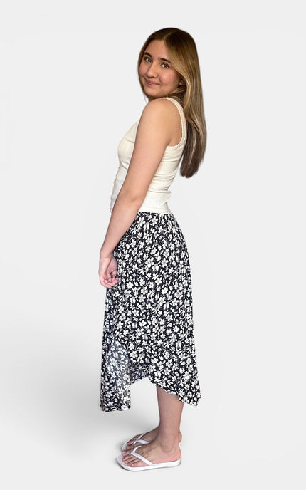 A woman modeling a black midi skirt with a white floral pattern, paired with a cream sleeveless top and white slide sandals, shown against a plain light gray background.