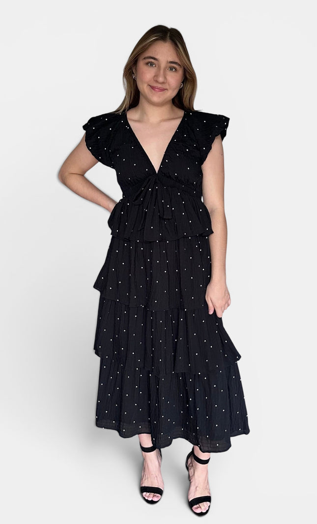 Black tiered midi dress with white polka dots, ruffled cap sleeves, and a tie waist, paired with black strappy heels against a light gray studio background.