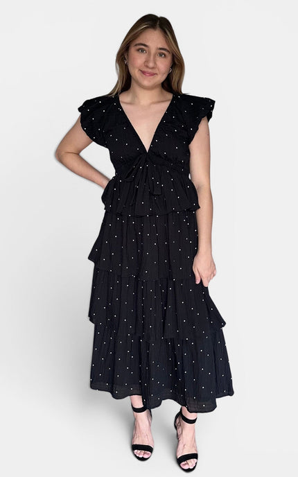 Black tiered midi dress with white polka dots, ruffled cap sleeves, and a tie waist, paired with black strappy heels against a light gray studio background.