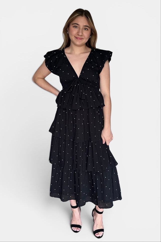 Black tiered midi dress with white polka dots, ruffled cap sleeves, and a tie waist, paired with black strappy heels against a light gray studio background.