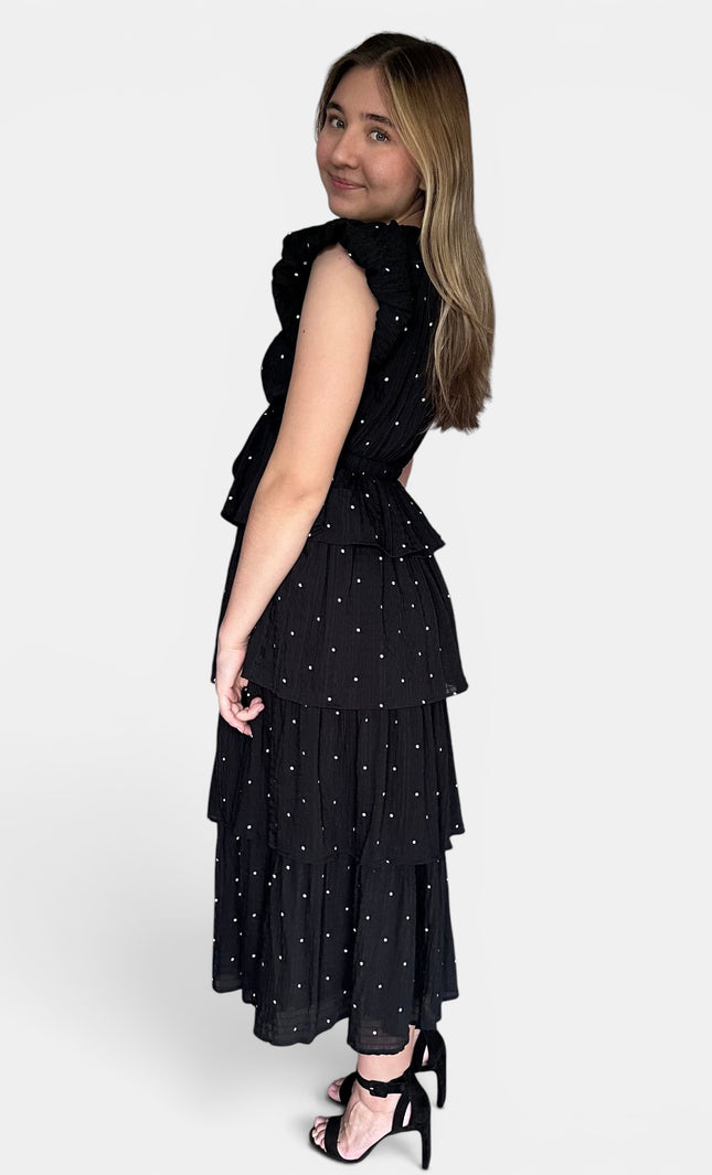 Sleeveless black midi dress with a white polka-dot pattern, featuring a ruffled high neckline and a tiered skirt, shown from a side-back angle against a light gray background.