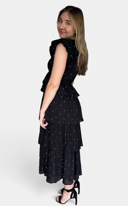 Sleeveless black midi dress with a white polka-dot pattern, featuring a ruffled high neckline and a tiered skirt, shown from a side-back angle against a light gray background.