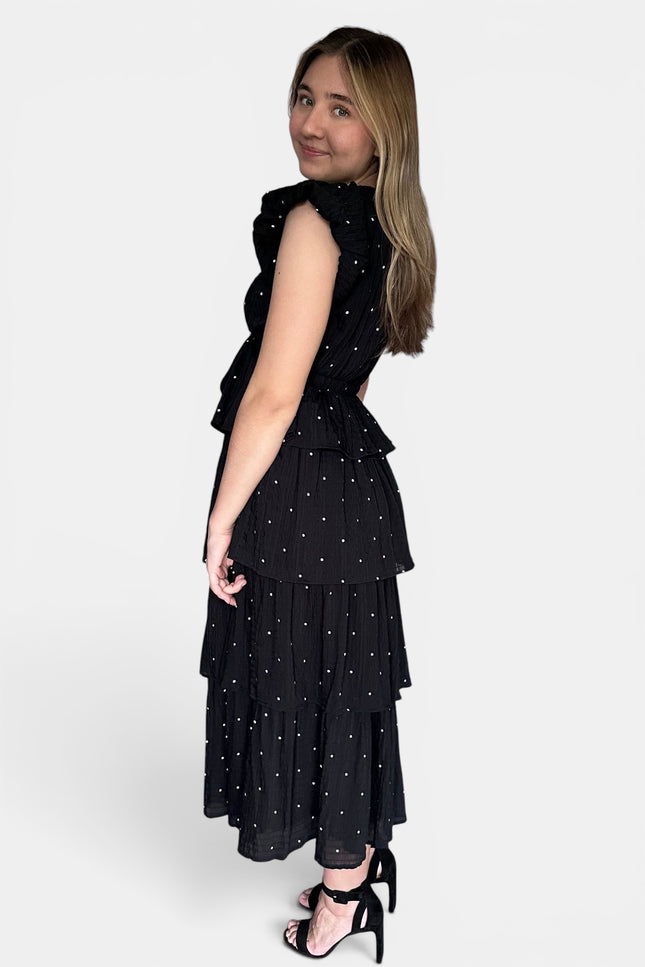 Sleeveless black midi dress with a white polka-dot pattern, featuring a ruffled high neckline and a tiered skirt, shown from a side-back angle against a light gray background.