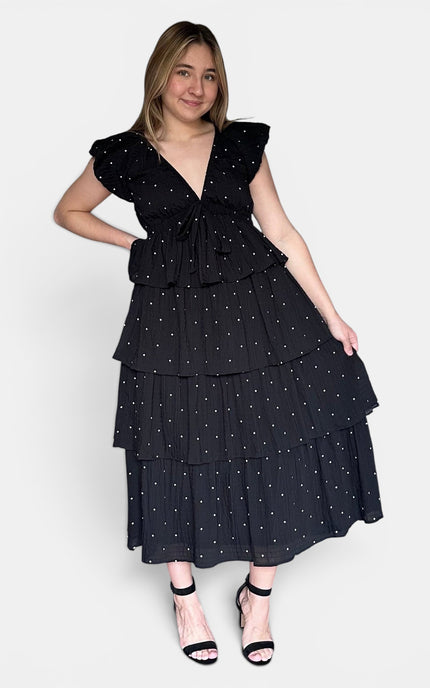 Black tiered midi dress with white polka dots, ruffled cap sleeves, and a tie waist, paired with black strappy heels against a light gray studio background.