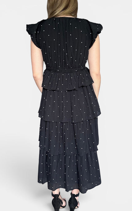 A woman shown from the back wearing a black tiered maxi dress with a white polka dot pattern and ruffled cap sleeves, paired with black heels, against a light gray studio background.