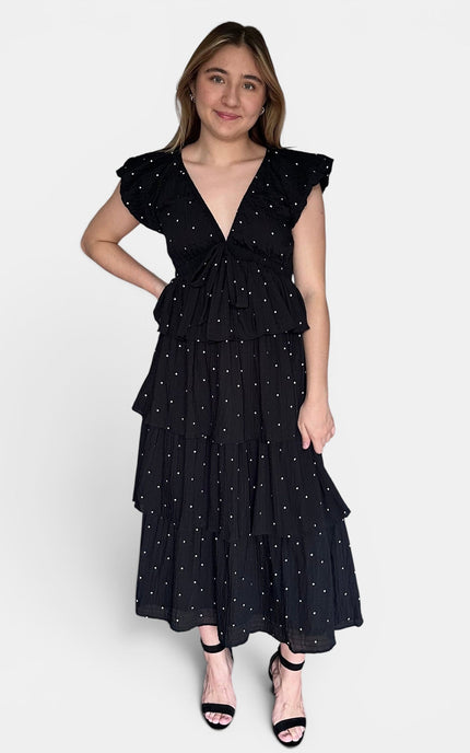 Black cotton tiered maxi dress with white polka dots, ruffled cap sleeves, and a ruffled waist tie, paired with black strappy heels against a light gray studio background.