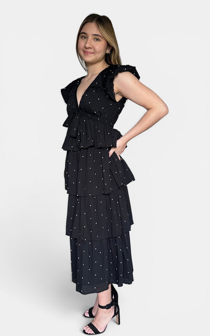 Black cotton tiered maxi dress with white polka dots and ruffled cap sleeves, paired with black strappy heels, against a light gray studio background.