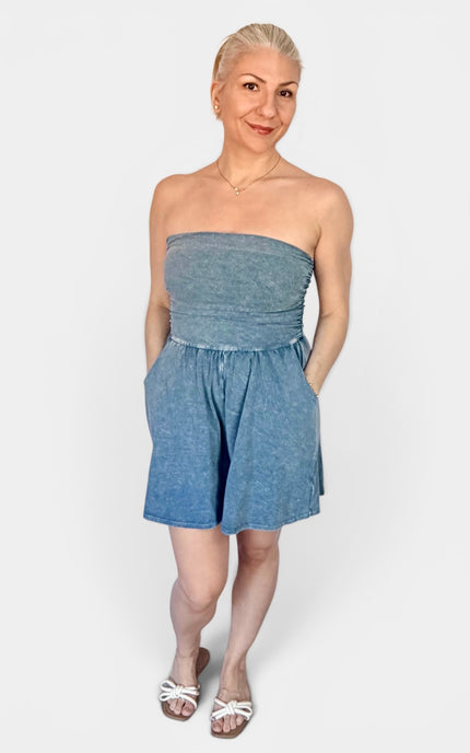 A woman modeling a strapless blue denim romper with a gathered waist and short flared legs, paired with white flat sandals, against a light gray studio background.