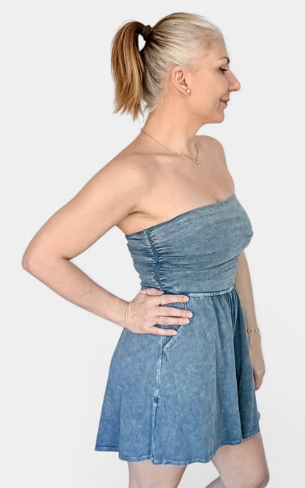 A woman modeling a strapless blue denim mini dress with a gathered bodice and flared skirt, shown in profile against a light gray studio background.