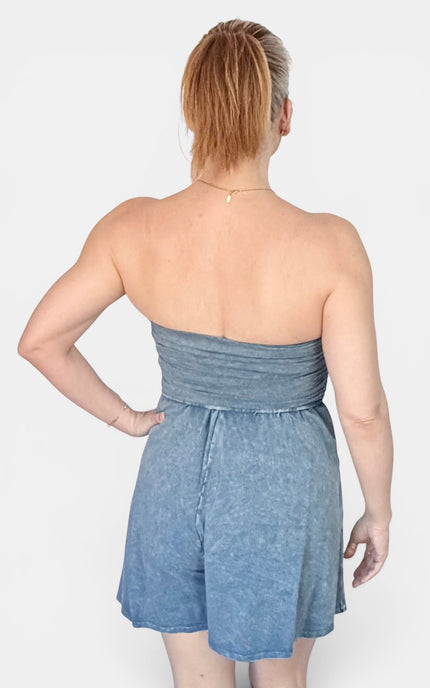 A woman shown from the back wearing a strapless blue denim romper with a ruched waistband and short flared legs, against a light gray studio background.
