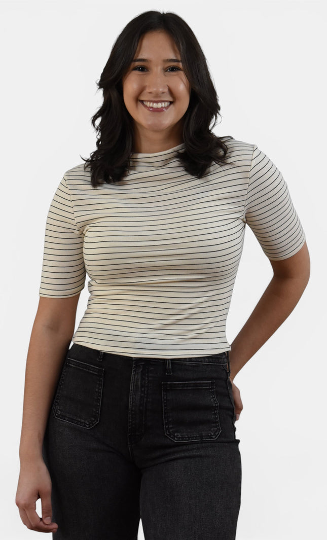 A woman wearing a beige short-sleeve crewneck top with thin dark horizontal stripes and black jeans, standing against a light gray studio background.