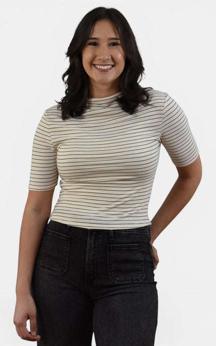 A woman wearing a beige short-sleeve crewneck top with thin dark horizontal stripes and black jeans, standing against a light gray studio background.