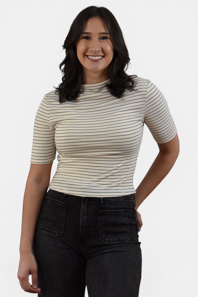 A woman wearing a beige short-sleeve crewneck top with thin dark horizontal stripes and black jeans, standing against a light gray studio background.