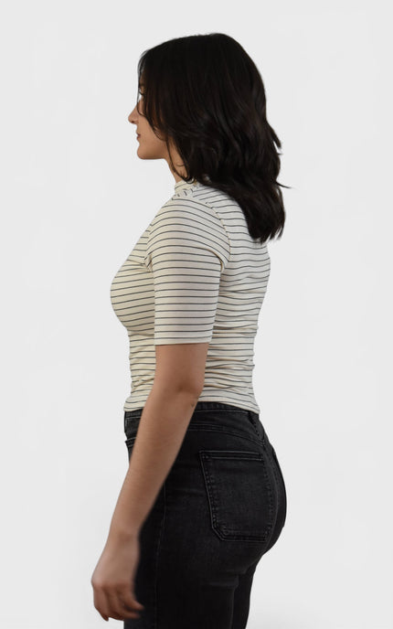 A woman shown from the side profile wearing a fitted short-sleeve top with thin horizontal dark stripes on a light beige or off-white base, paired with dark jeans, against a plain light gray studio background.