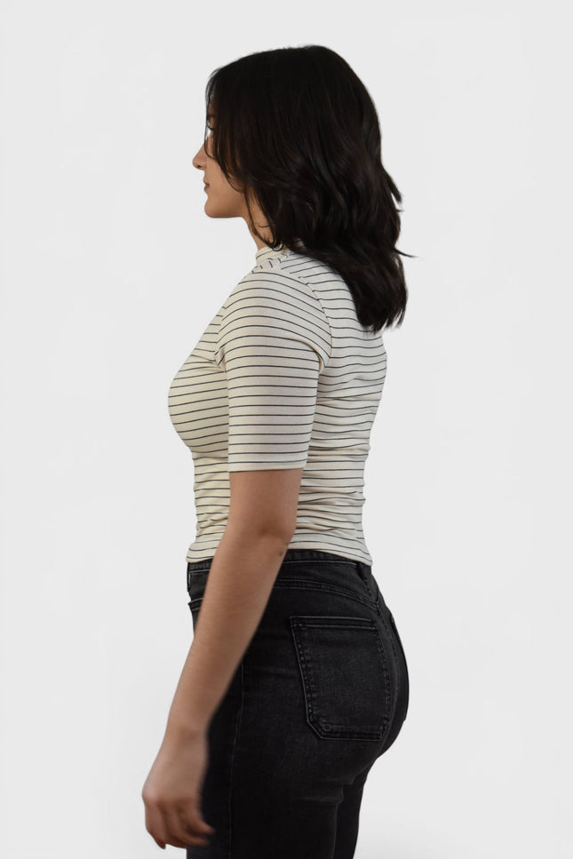 A woman shown from the side profile wearing a fitted short-sleeve top with thin horizontal dark stripes on a light beige or off-white base, paired with dark jeans, against a plain light gray studio background.
