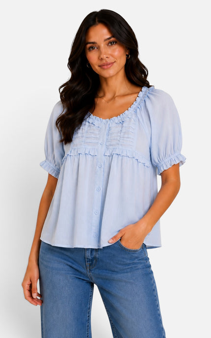 A woman modeling a light blue short-sleeve blouse with ruffled trim and button details, paired with blue jeans, against a plain white studio background.