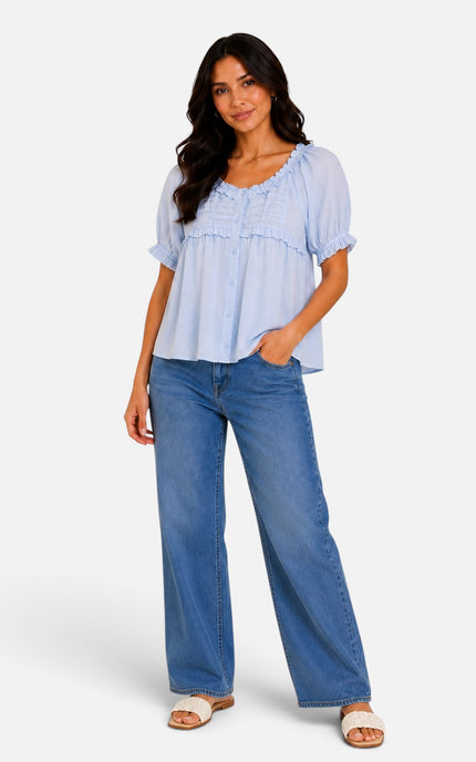 A woman modeling a light blue short-sleeve blouse with ruffled trim and blue wide-leg jeans against a plain light gray studio background.
