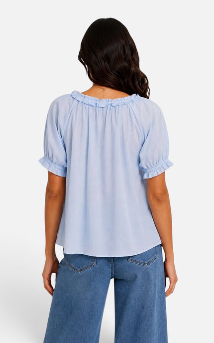 A woman viewed from the back wearing a light blue short-sleeve blouse with ruffled trim at the neckline and cuffs, paired with blue jeans, against a plain light gray studio background.