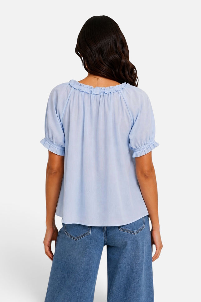A woman viewed from the back wearing a light blue short-sleeve blouse with ruffled trim at the neckline and cuffs, paired with blue jeans, against a plain light gray studio background.
