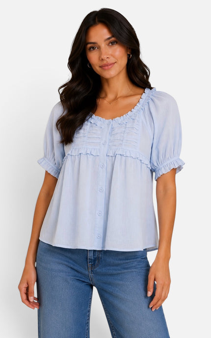 A woman modeling a light blue short-sleeve blouse with ruffled trim and button details, paired with blue jeans, against a plain white studio background.