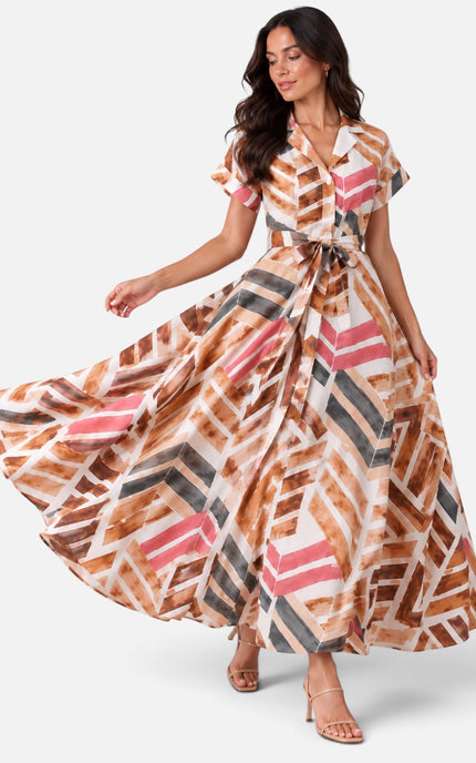 A woman modeling a maxi dress with a geometric brushstroke pattern in brown, pink, gray, and white, posed against a light gray studio background.
