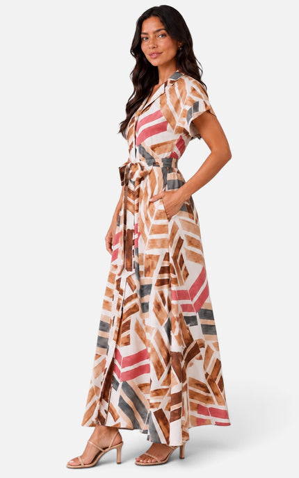 A woman modeling a long maxi dress with a geometric abstract print in brown, pink, gray, and white, paired with beige heeled sandals against a light gray studio background.
