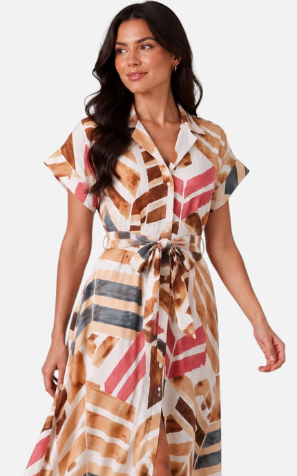 A woman modeling a short-sleeve maxi dress with a geometric abstract print in brown, beige, pink, and gray on a white background, paired with beige strappy high-heeled sandals.