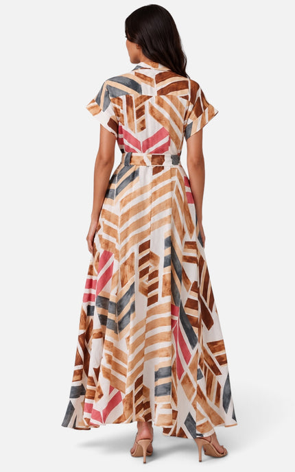 A woman viewed from the back wearing a short-sleeve maxi dress with a geometric abstract print in brown, beige, pink, and gray on a white base, paired with beige high heels, set against a light gray studio background.