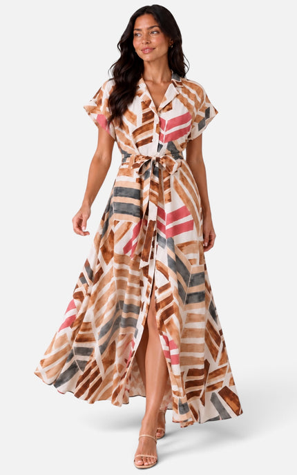 A woman modeling a long maxi dress with a geometric abstract print in brown, beige, pink, and gray, set against a plain light gray studio background.