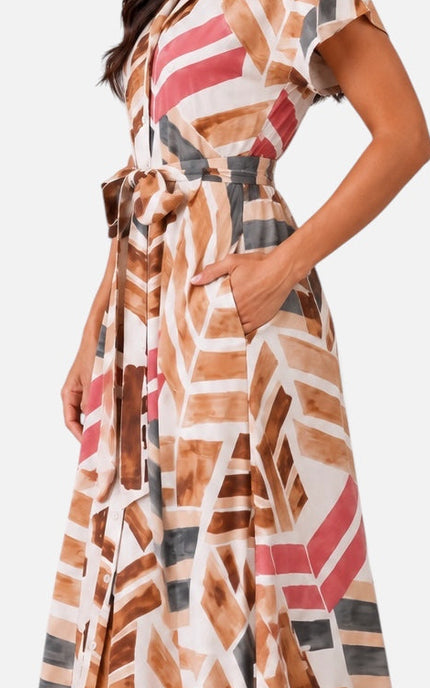 A woman modeling a long maxi dress with a geometric abstract print in brown, pink, gray, and white, paired with beige heeled sandals against a light gray studio background.