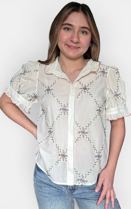 A woman wearing a white short-sleeve button-up blouse with a delicate floral and geometric embroidered pattern, paired with blue jeans, against a plain light gray background.