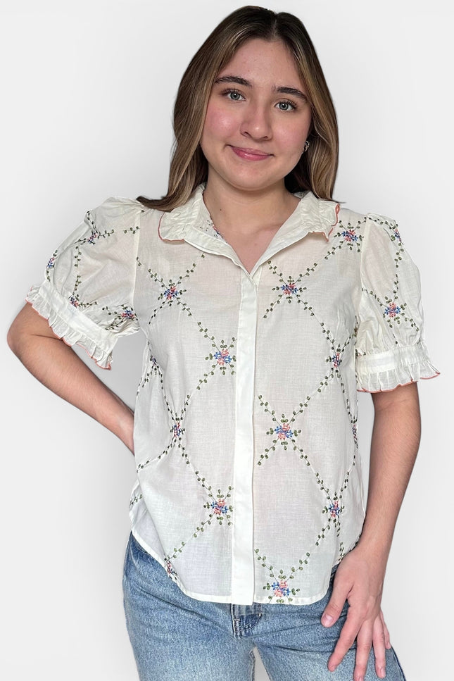 A woman wearing a white short-sleeve button-up blouse with a delicate floral and geometric embroidered pattern, paired with blue jeans, against a plain light gray background.
